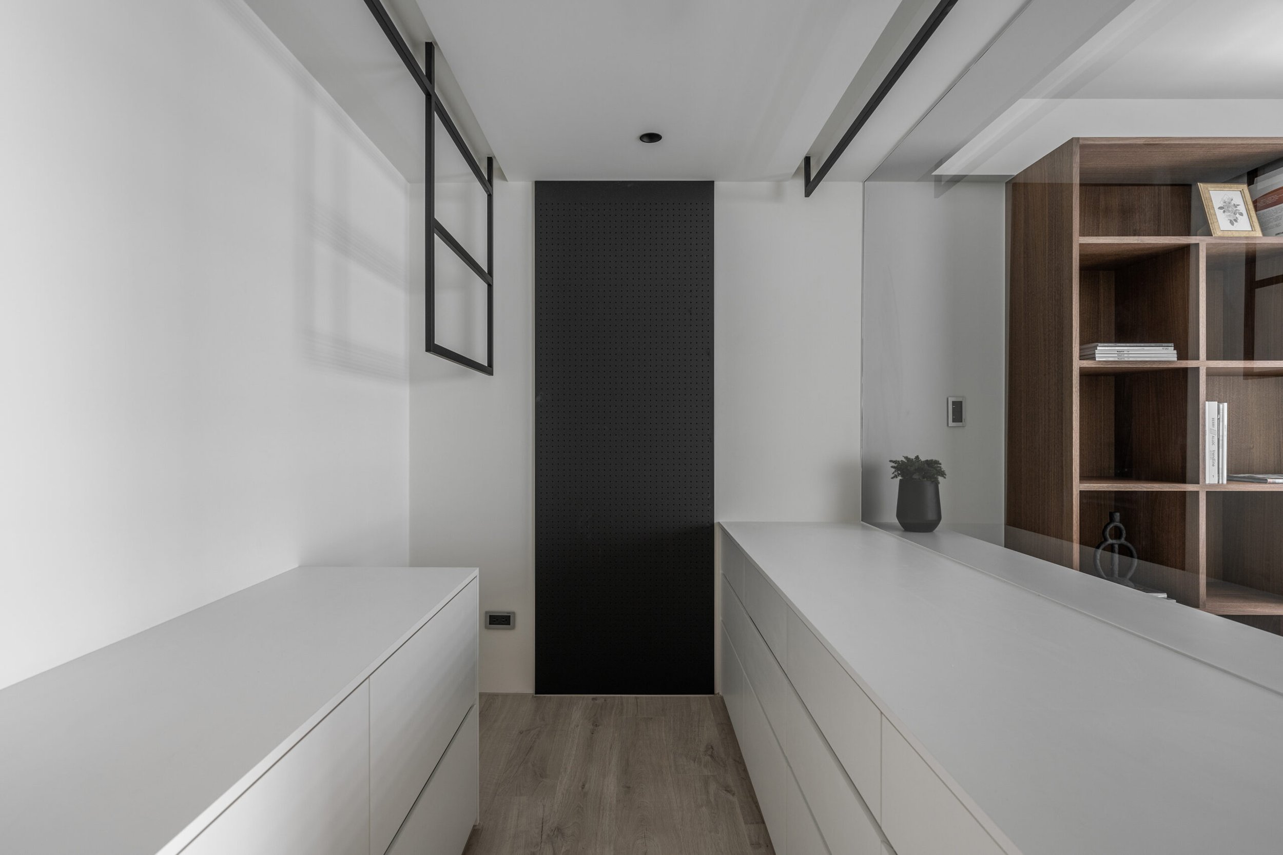 Modern hallway with white cabinets on both sides, black pegboard wall at the end, wooden bookshelf with framed photo and books, small potted plant, and wood flooring.
