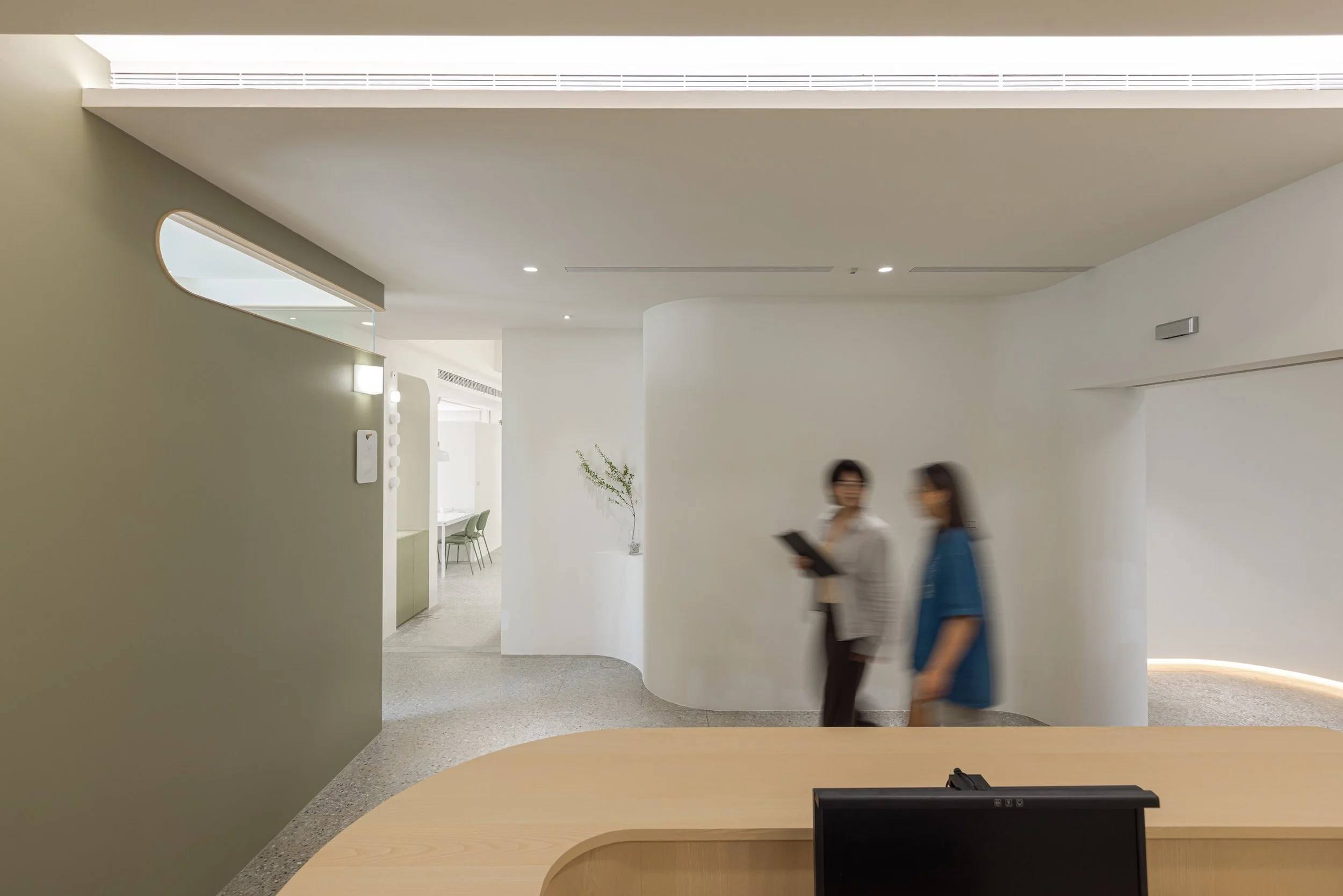 Modern interior space with white walls, curved architecture, and two people walking. The area has minimalistic design with a high ceiling, circular cutouts, and simple furniture.