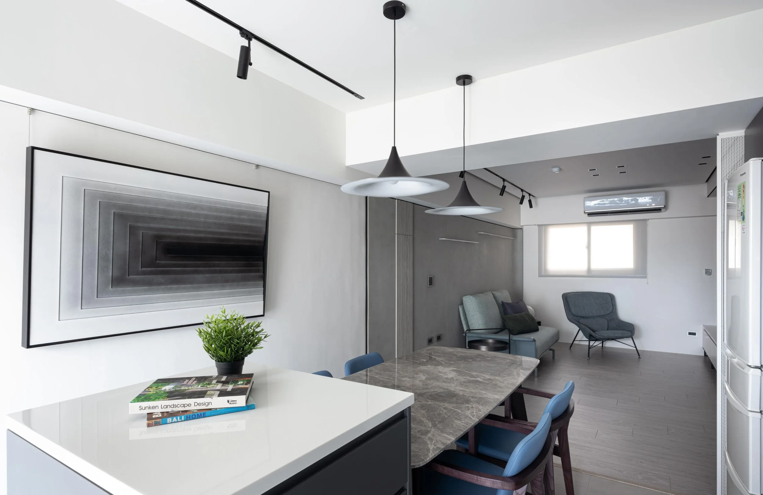 Modern living room with white walls and ceiling, black track lighting, two black pendant lights, wall-mounted air conditioner, a large abstract black and white art piece, a gray sofa, two gray armchairs, a marble dining table with blue chairs, a small potted plant, and a couple of magazines on a white kitchen island.