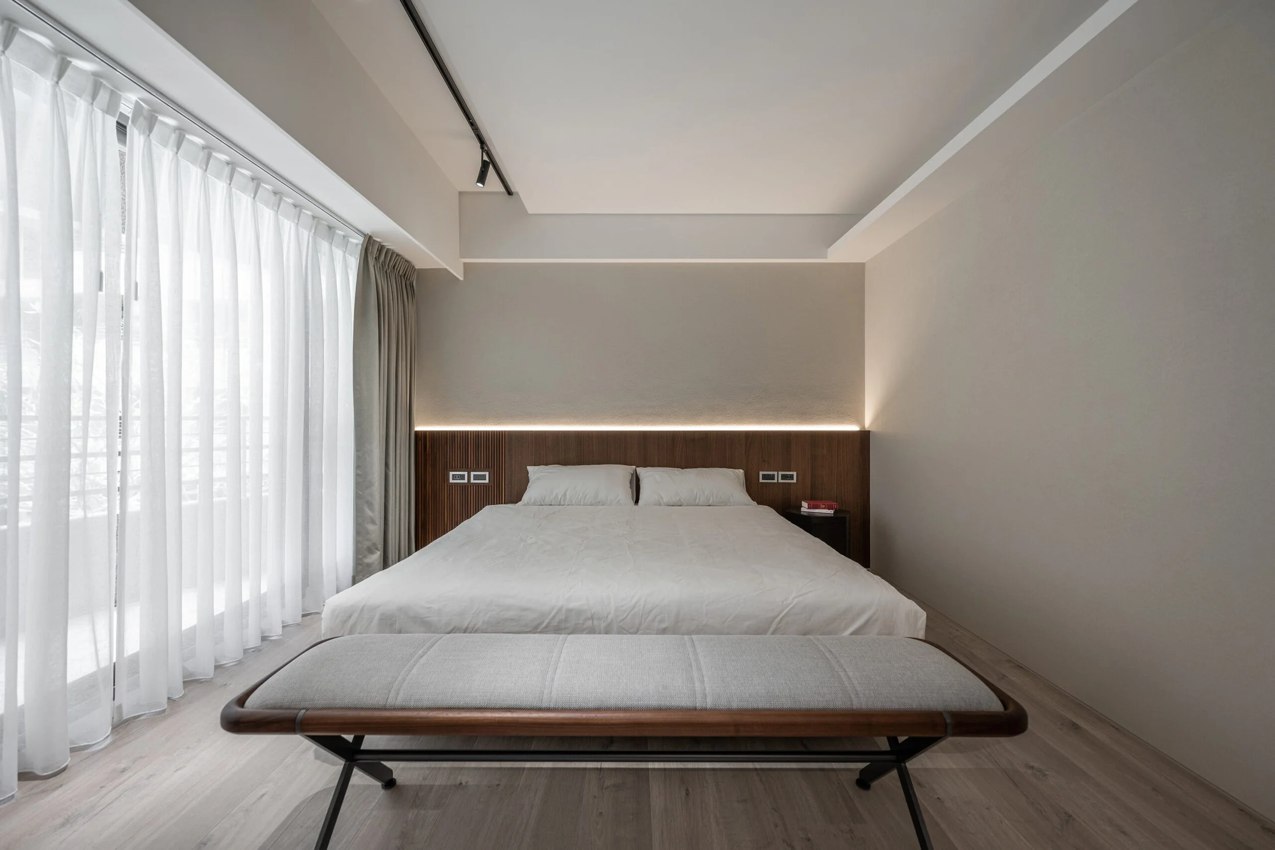 Modern bedroom with large bed, wooden headboard, white bedding, side table, and tall window with sheer and blackout curtains, with a gray lounge chair at the foot of the bed.