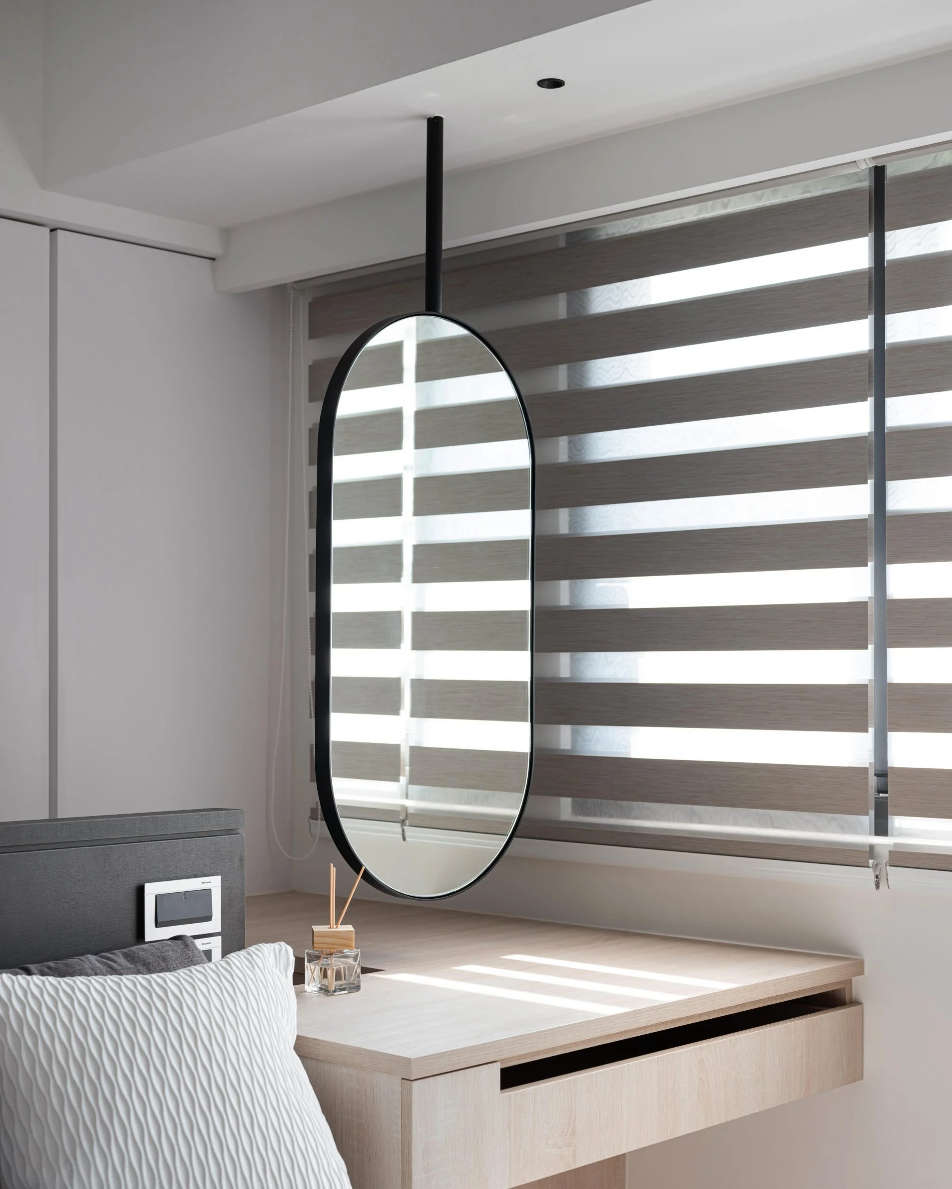 A minimalist bedroom corner with a light wood desk, a large pillow with a textured white cover, a small glass container with reed diffusers, a mounted circular mirror with a black frame, a window with striped blinds allowing sunlight to enter, and a grey speaker or electronic device on the wall.