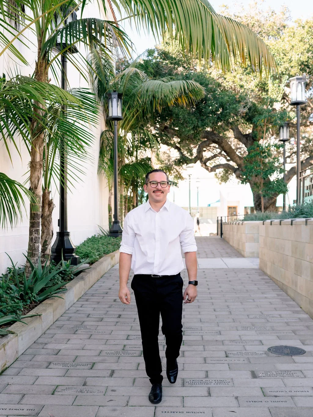 A man in a white shirt and black pants walking outside on a brick path, surrounded by green palm trees and large trees, with light posts and a fence in the background.