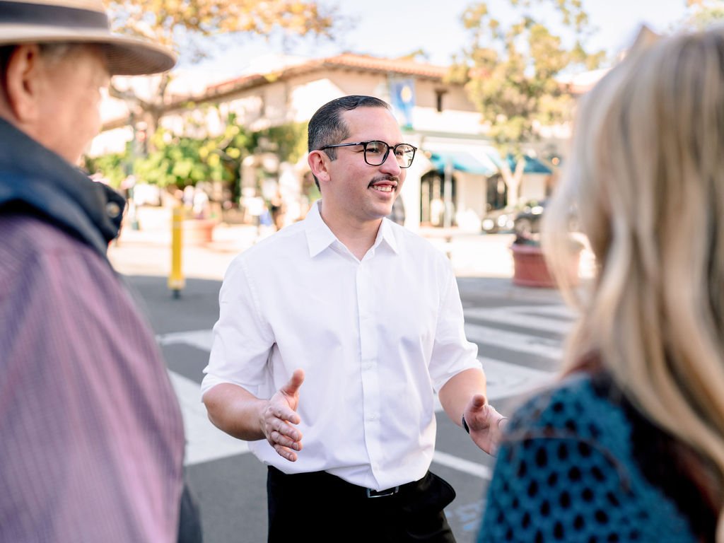 A man in a white shirt with glasses talking to two women on a city street during the day.