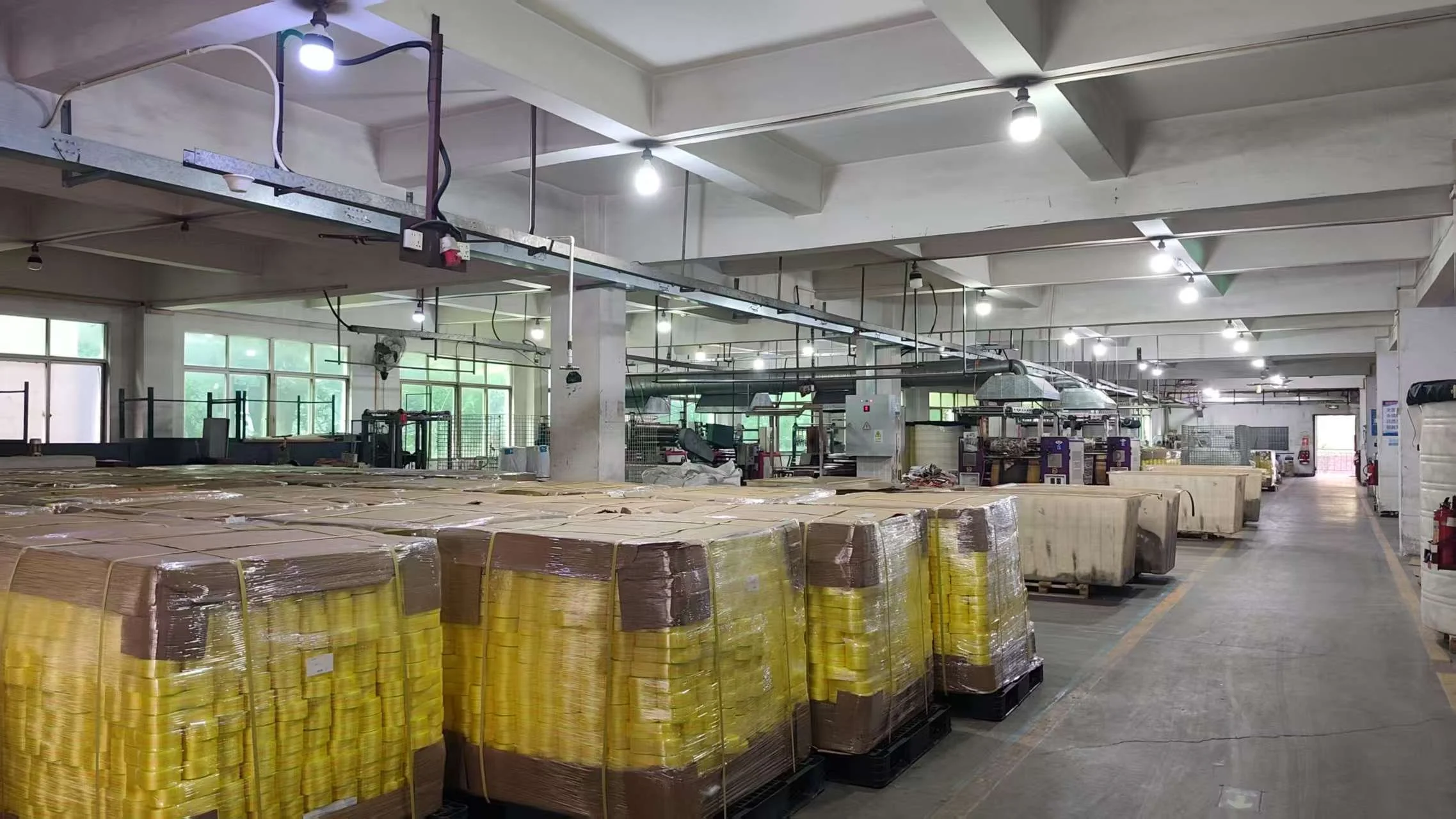 Warehouse or factory interior with pallets of packaged goods and industrial equipment, ceiling lights, windows, and a door in the background.