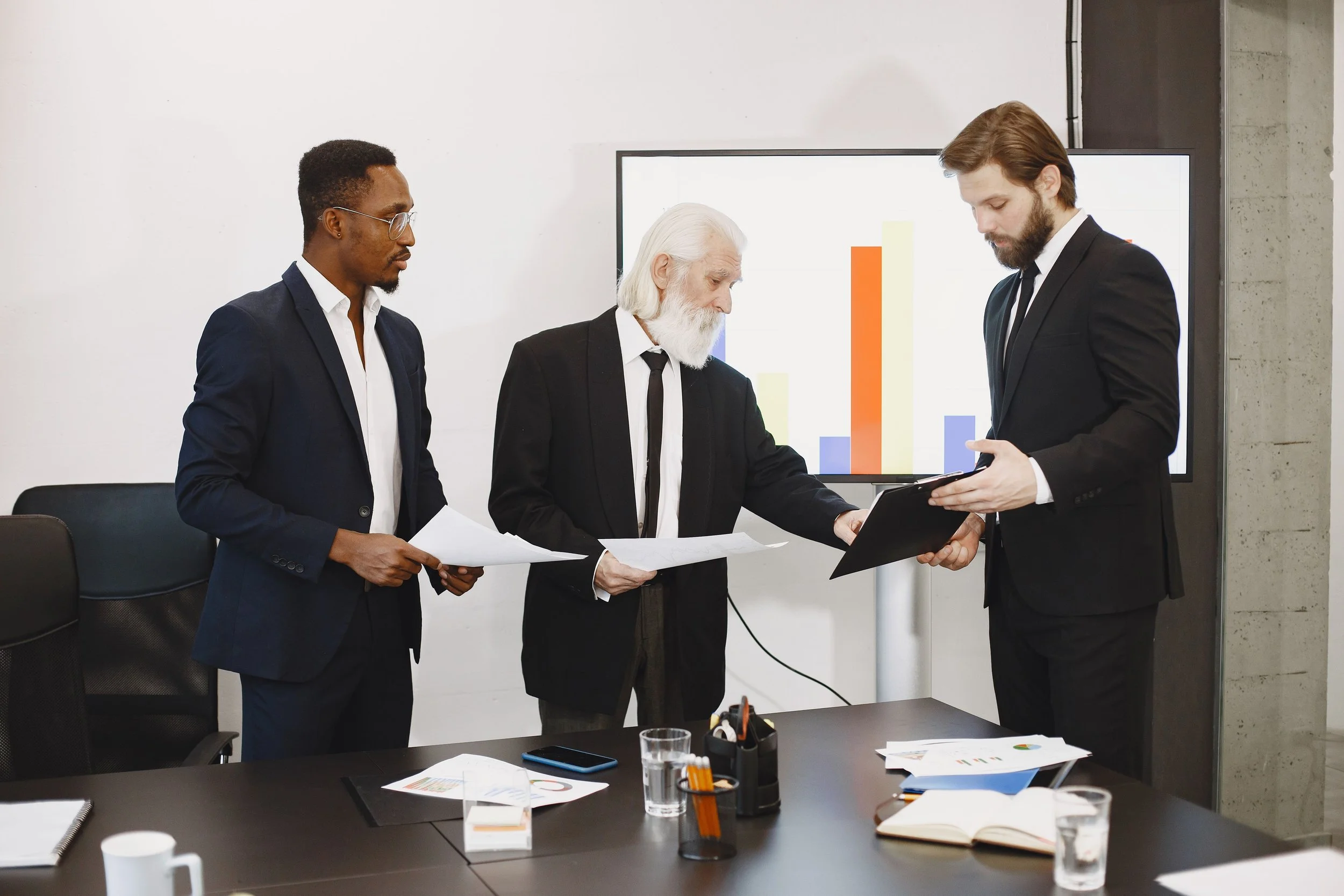 Three men in suits are gathered in a business meeting room. They are reviewing documents, standing near a table with office supplies and beverages. A monitor behind them displays colorful bar charts.