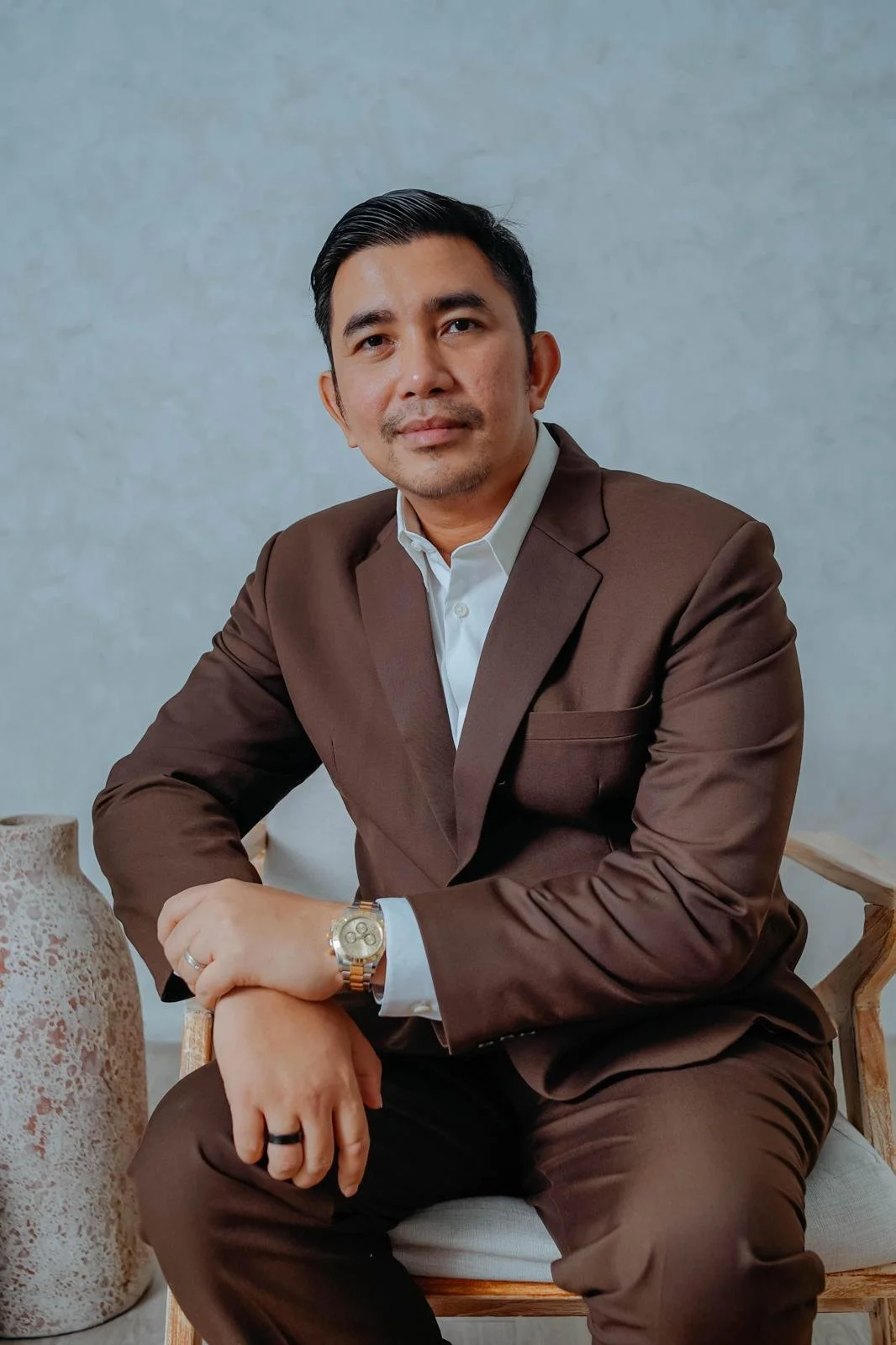 A man in a brown suit with a white shirt, sitting on a wooden chair, wearing a watch and ring, leaning on his arm, against a gray wall.