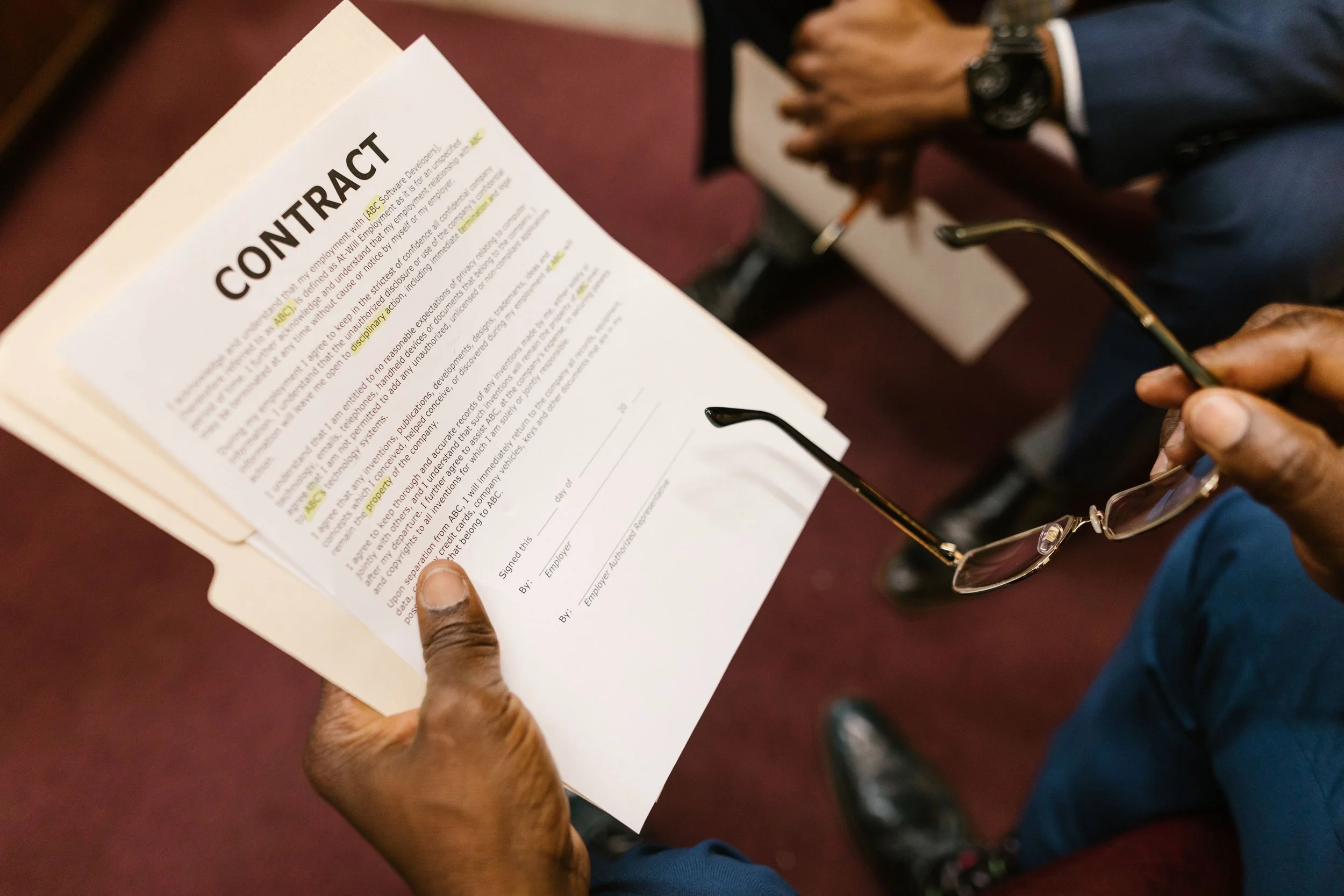 Close-up of a person holding a contract document with highlighted text, and another person in the background taking notes or signing on a clipboard.