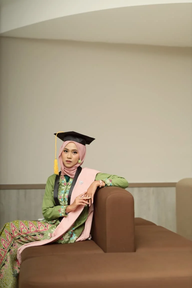 A woman in a graduation gown and cap sitting on a brown couch in a room with plain walls.