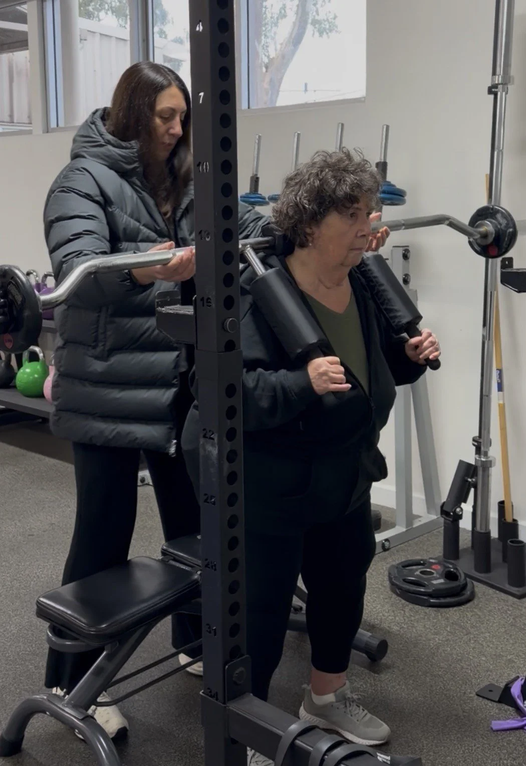 An older woman is doing a squat exercise with a barbell on her shoulders, assisted by a trainer behind her in a gym.