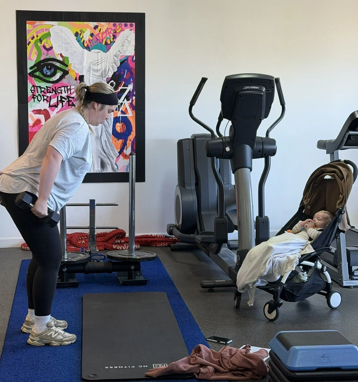 A woman is doing a workout with dumbbells in a home gym, while a baby in a stroller is resting or sleeping nearby. There is exercise equipment including a treadmill, a rowing machine, and weights, with a colorful artistic poster on the wall in the background.