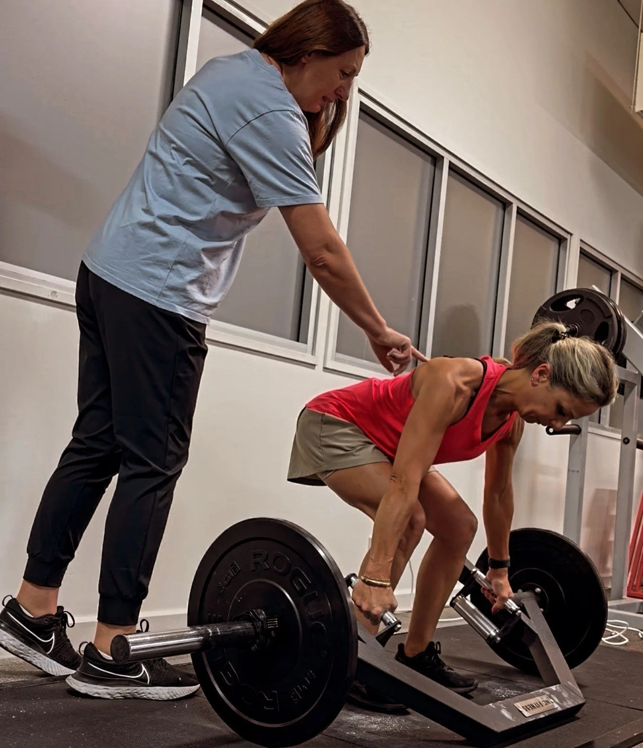 A woman performing a deadlift exercise with weights on a gym platform, assisted by a trainer standing behind her, in an indoor gym.