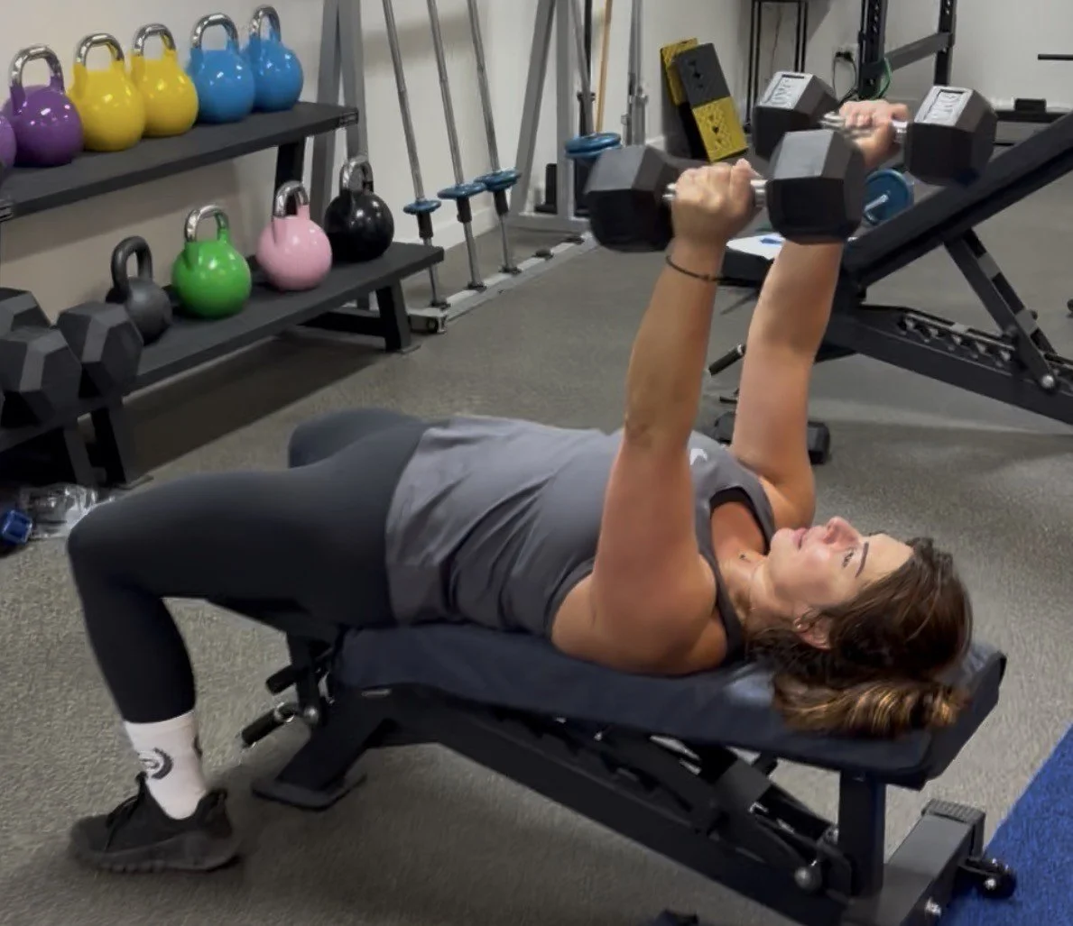 A woman lying on a workout bench lifting dumbbells in a gym with colorful kettlebells and training equipment in the background.