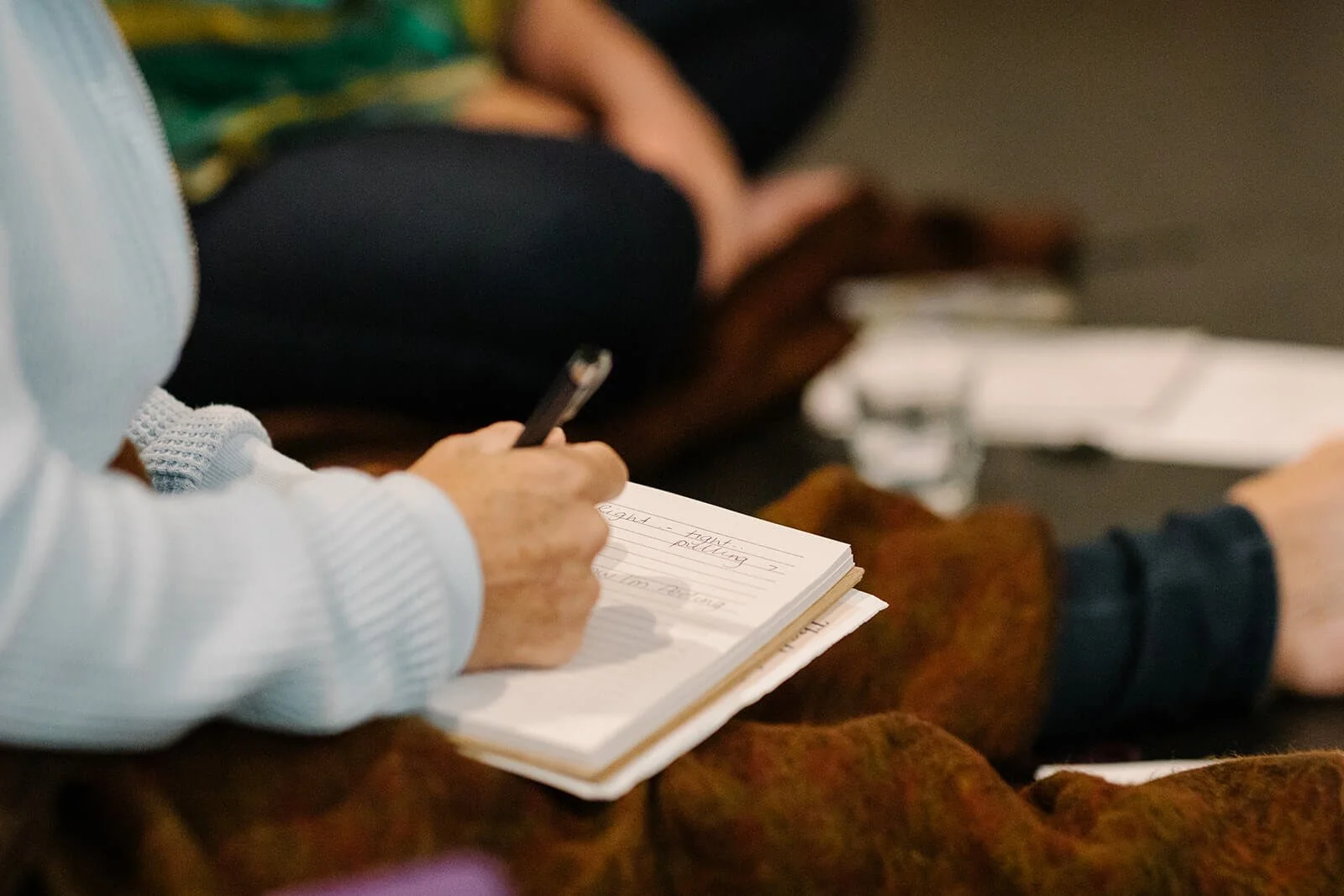 A person writing in a notebook with a pen, seated on a brown and orange patterned fabric, with other people sitting nearby.