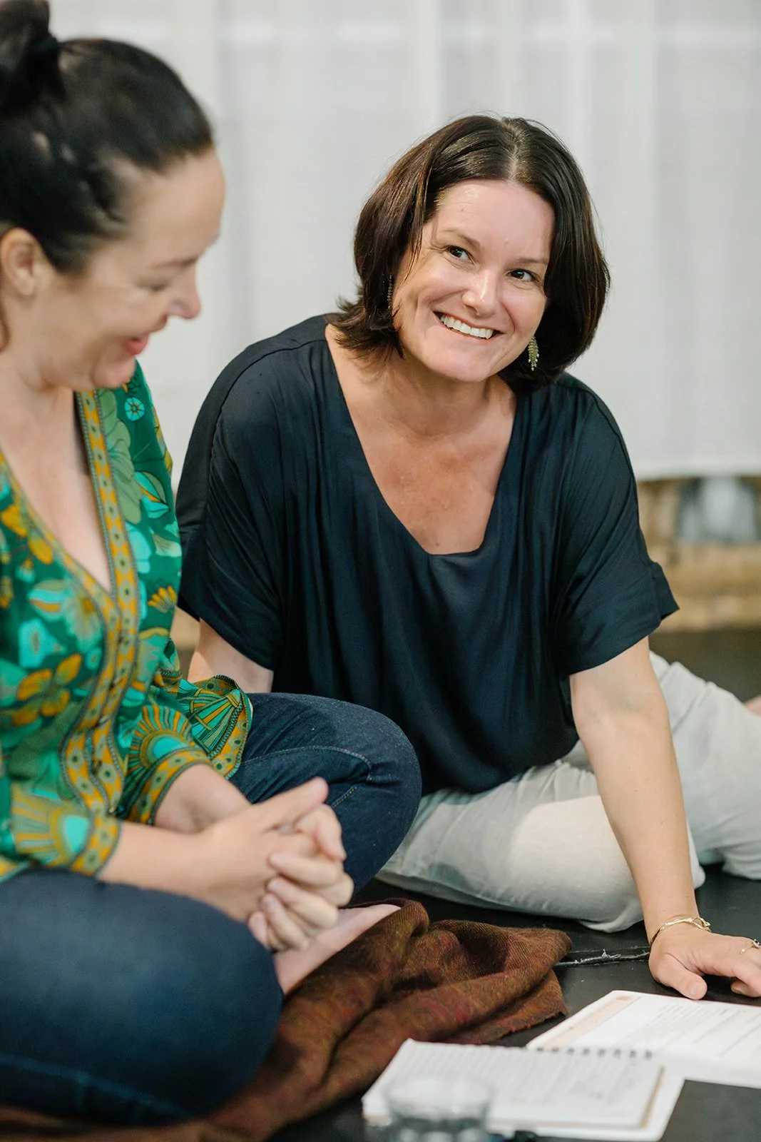 Two women sitting and smiling during a Divine EFT & Wllness discussion, with open books and papers in front of them.