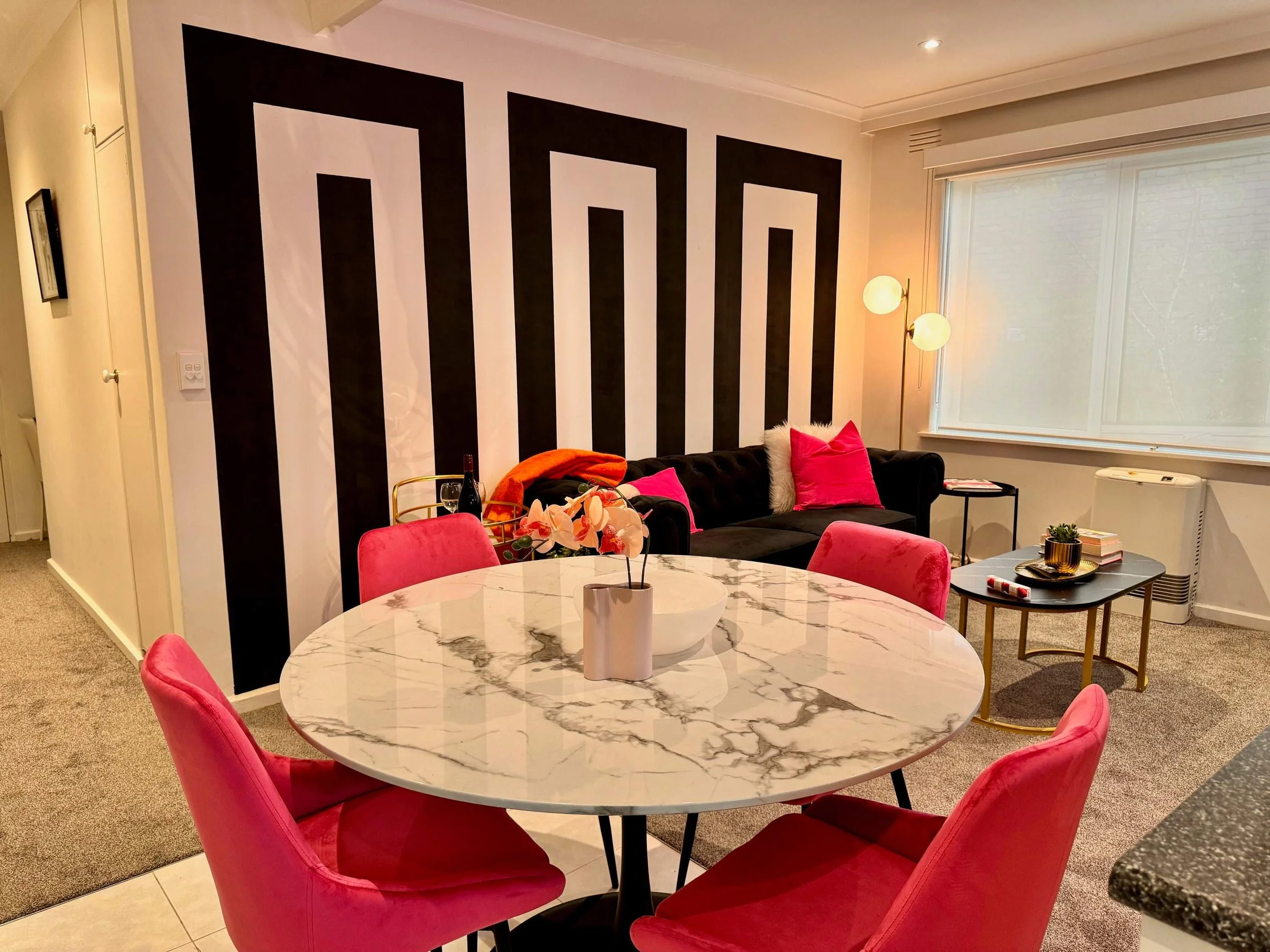A living room featuring a marble dining table with pink chairs, a black couch with pink and white pillows, a tall floor lamp, and a window with a view of trees outside. There is a feature wall with black and white vertical stripes, and a small side t