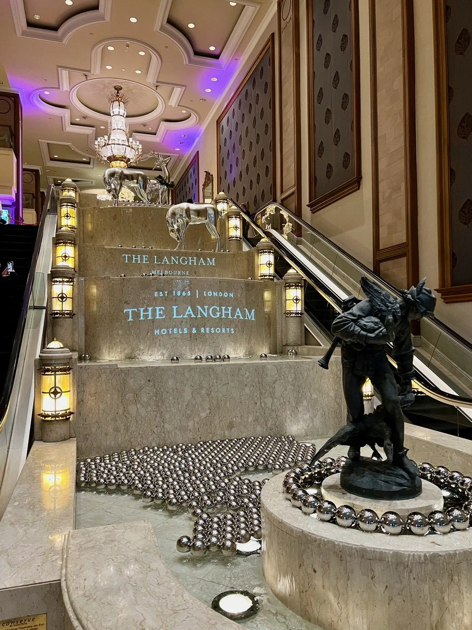 Interior lobby of The Langham hotel in Melbourne featuring a staircase with silver ornaments, sculptures of reindeer and a wolf, decorative lighting, and purple and warm wall lighting.