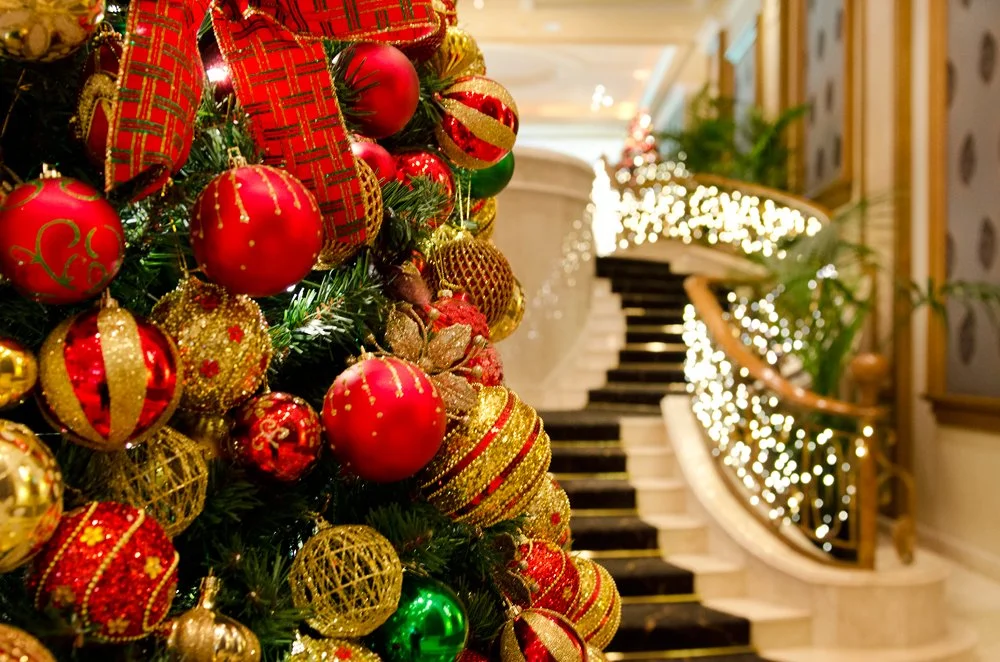 Close-up of decorated Christmas tree with red, gold, and green ornaments in a hotel or mall lobby with lit balustrades and staircase in the background.