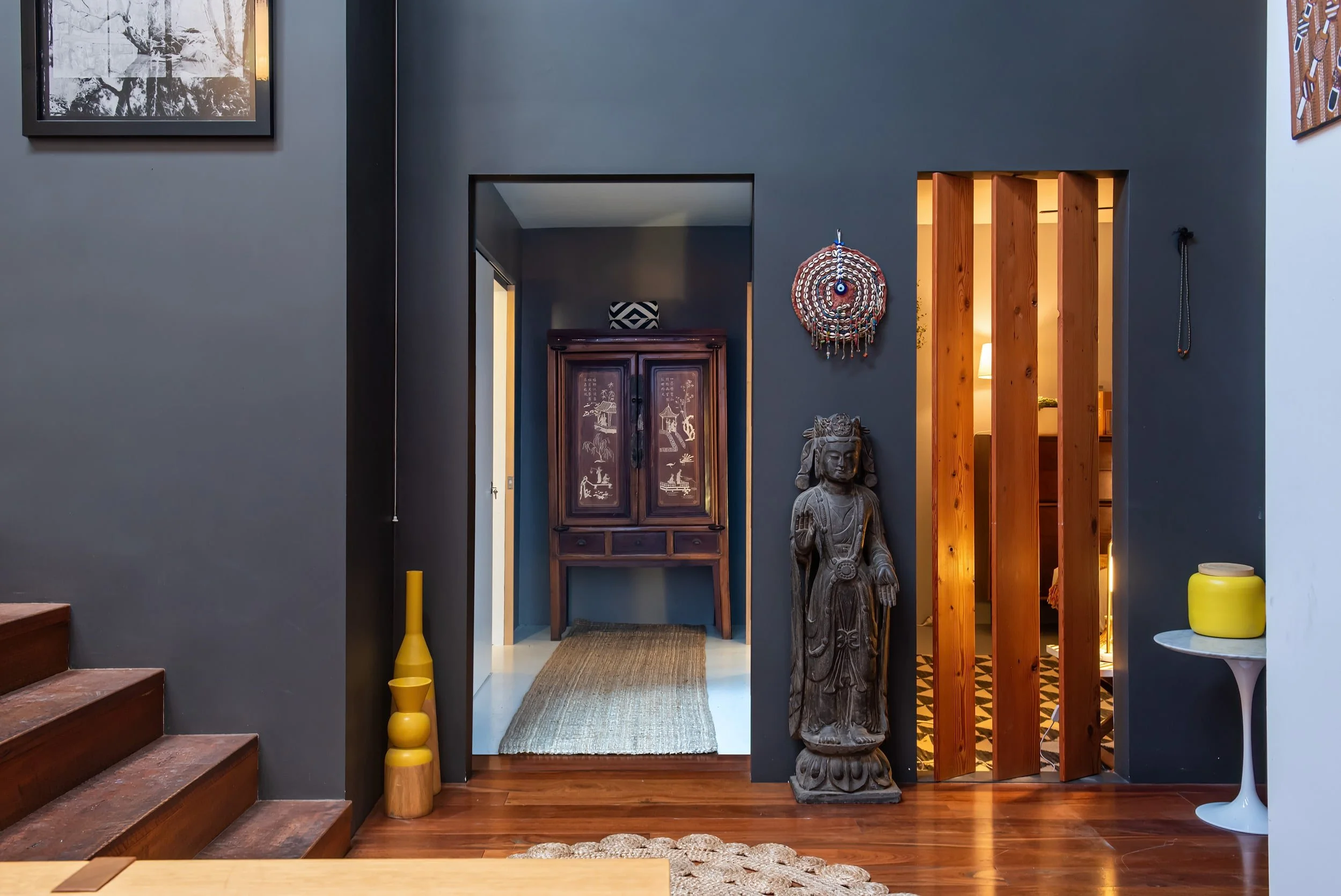 Indoor decor with blue walls, wooden staircase, vintage wooden cabinet, tribal statue, colorful wall hanging, yellow vases, and a side table with a yellow jar.