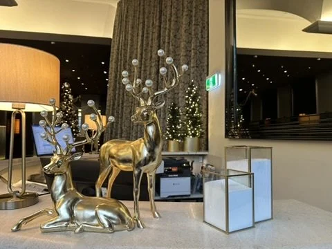 Decorative gold reindeer statues with white spherical ornaments on antlers, placed on a table in a decorated interior space with warm lighting and Christmas trees in the background.