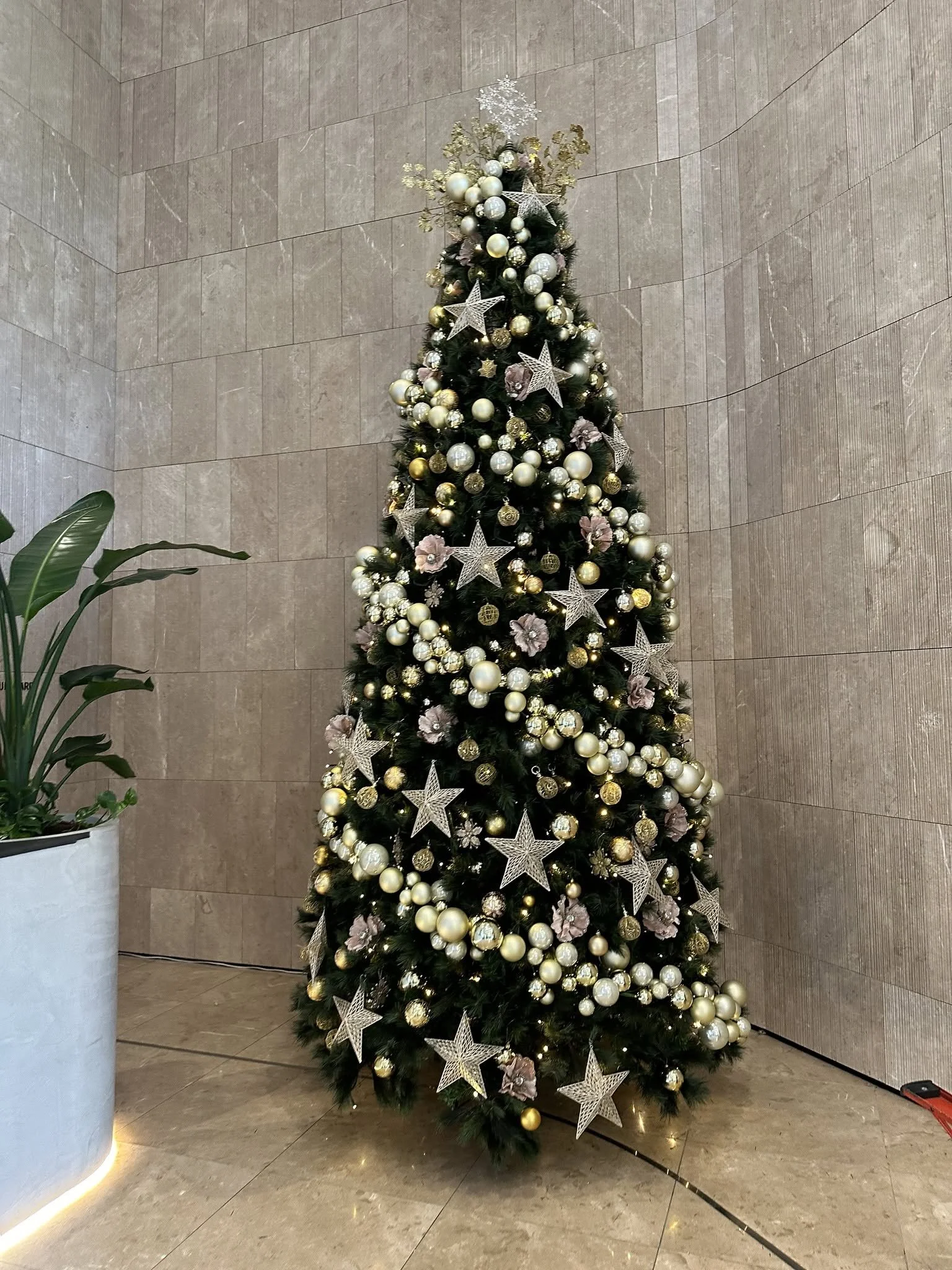A decorated Christmas tree with white, gold, and pink ornaments, stars, and flowers, standing against a tiled wall in an indoor setting, with a potted plant nearby.