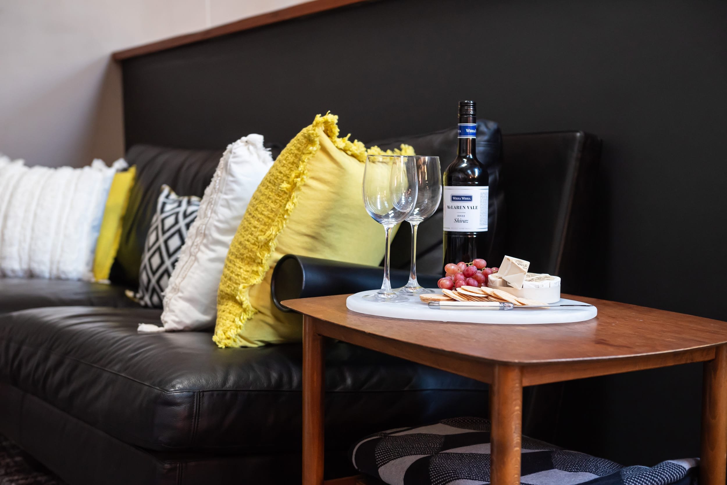 A black leather couch with colorful pillows behind a wooden coffee table displaying a wine bottle, two wine glasses, grapes, cheese, and crackers, with a dark wall in the background.