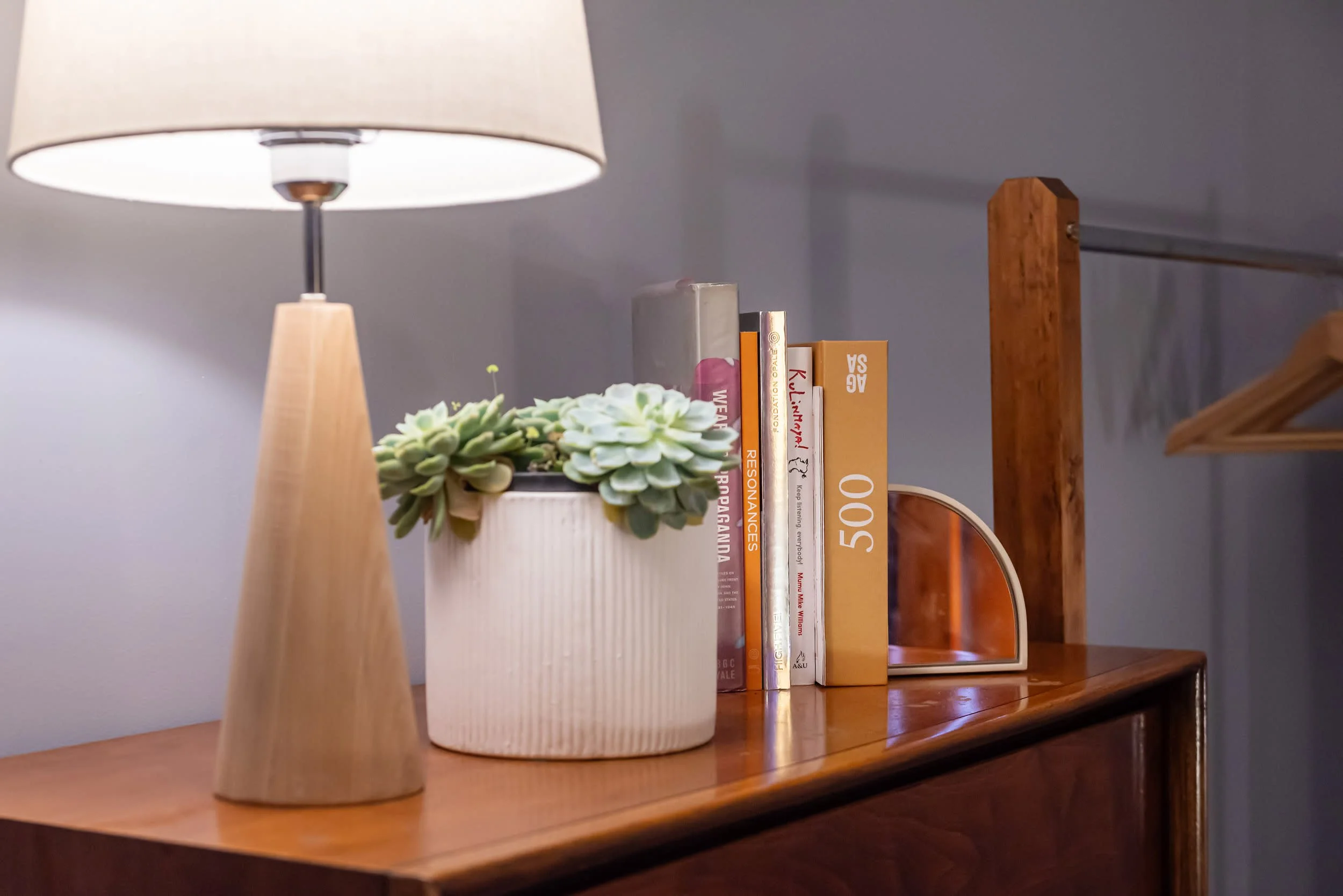 A wooden table with a beige lamp, a potted succulent plant, a small mirror, and a row of books on a wooden shelf against a gray wall.