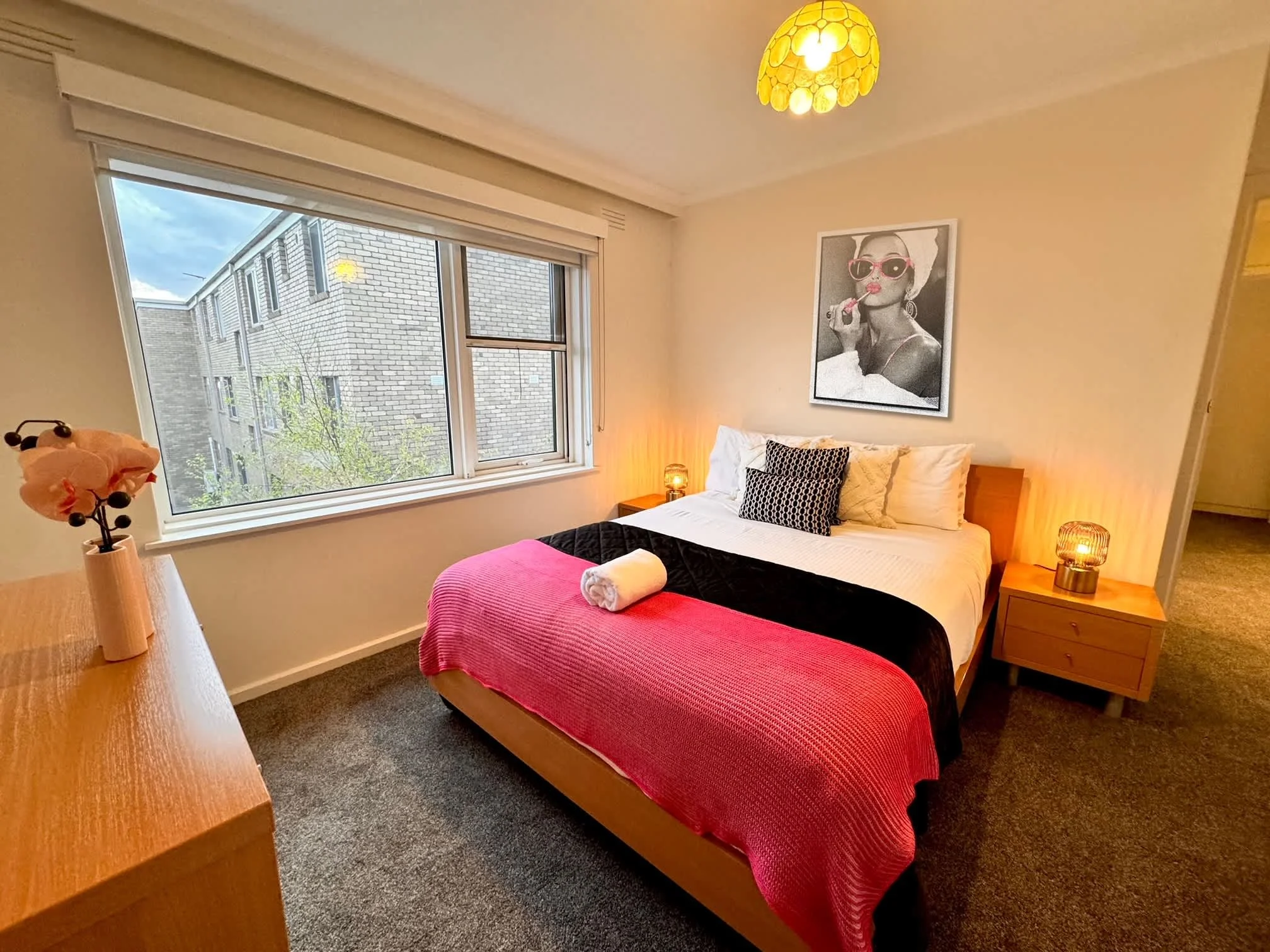 A bedroom with a large window showing a brick building and cloudy sky outside. The bed has white sheets with a black and white patterned pillow and a rolled white towel at the foot. There are two wooden nightstands with small lamps, and artwork of a 