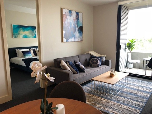 Living room with gray sofa, decorative pillows, abstract wall art, and a patterned rug. Bedroom visible through open door with bed and wall art, and a balcony with outdoor seating and greenery.