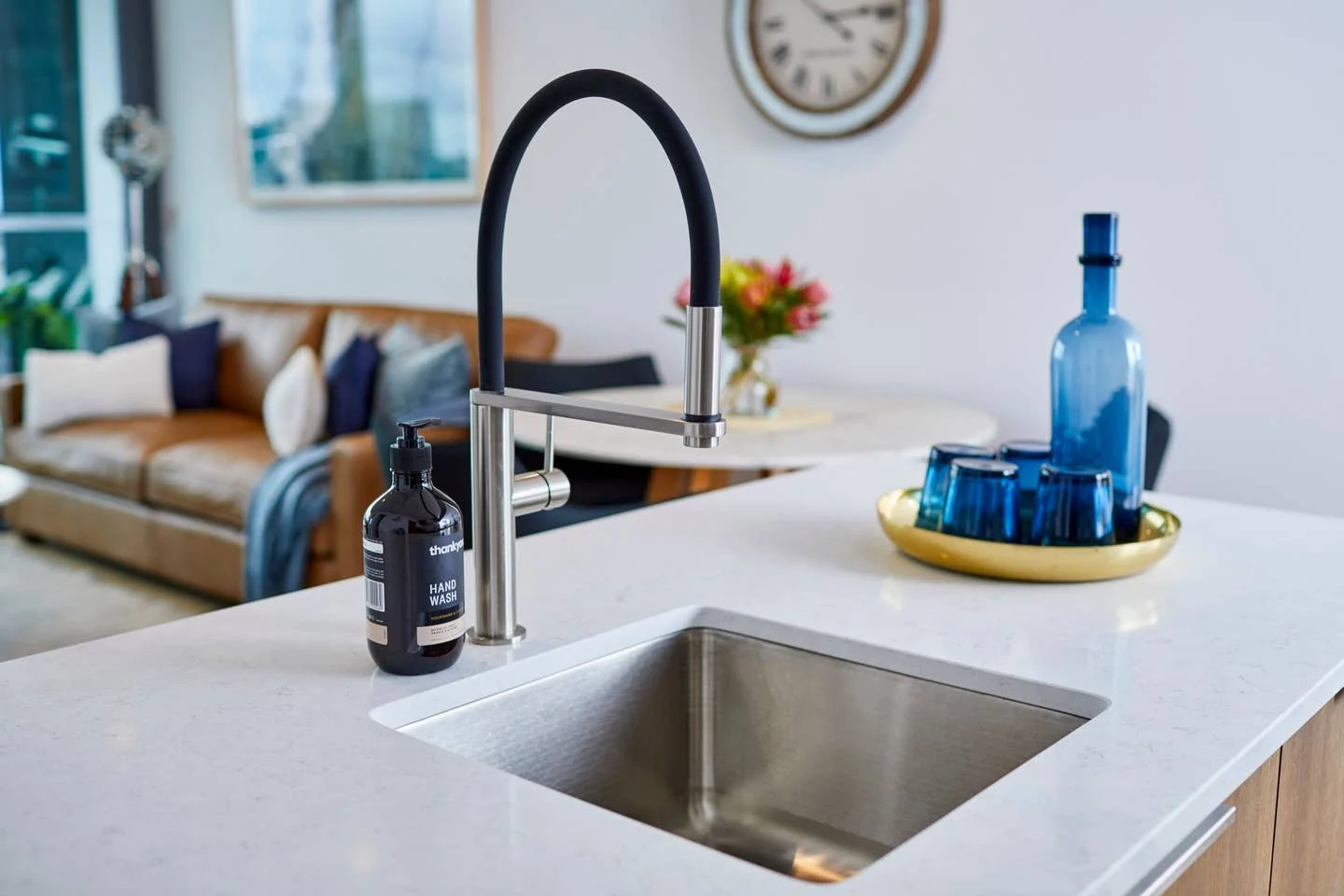 Modern kitchen with a black and stainless steel faucet, hand wash soap dispenser, blue glassware, and a gold tray, with a living room in the background.