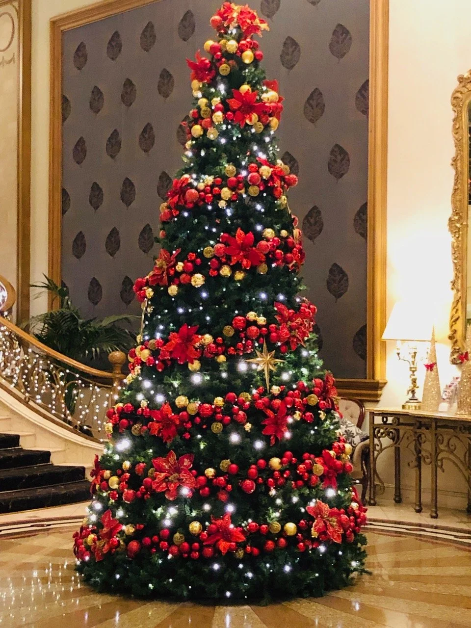 Decorated Christmas tree at the Langham Hotel Melbourne with red and gold ornaments, poinsettia flowers, and white lights in a lavish interior with a staircase, plant, and a side table with a lamp in the background.
