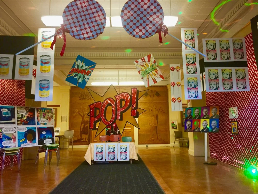 Decorative pop art-themed display with comic book style speech bubbles and images, featuring a large sign that says 'POP!', and images of soup cans, portraits of people, and vintage phones in a room decorated with red polka dot patterns.
