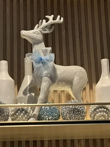 Decorative reindeer ornament with a blue bow on its neck, displayed on a glass shelf with white and blue Christmas ornaments and vases in the background.