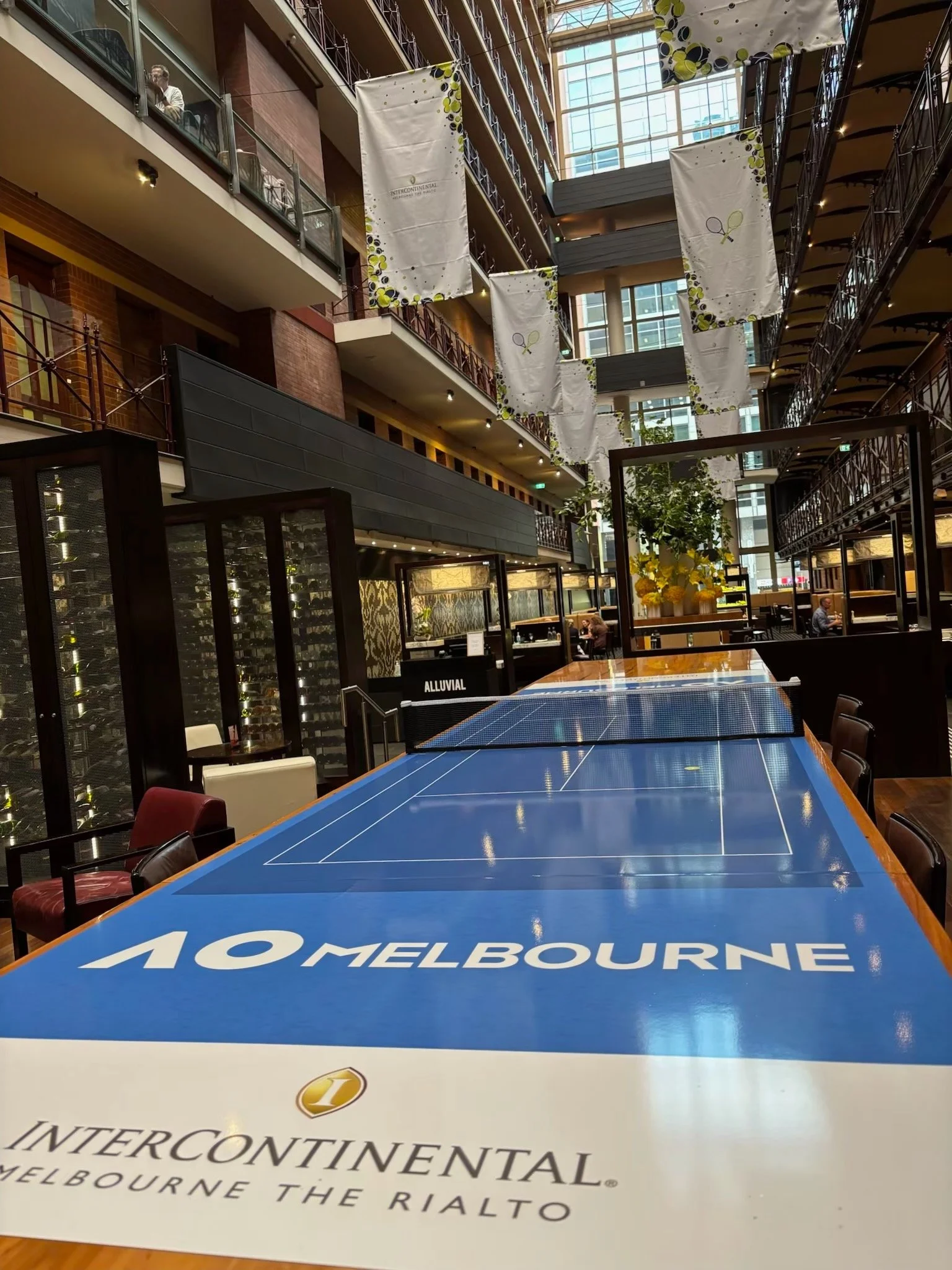 Indoor venue with multiple stories, large windows, and banners hanging from the ceiling, featuring a ping pong table with the words 'MELBOURNE THE RIALTO' on its side, and some people seated and walking around.