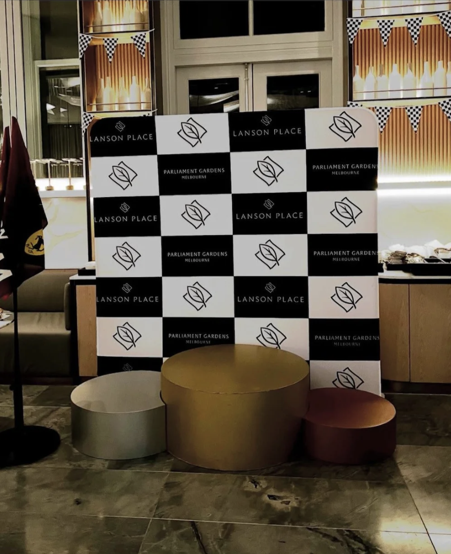 Event backdrop with logos of Lanson Place and Parliament Gardens Melbourne, two gold and bronze circular tables, in a decorated indoor space.