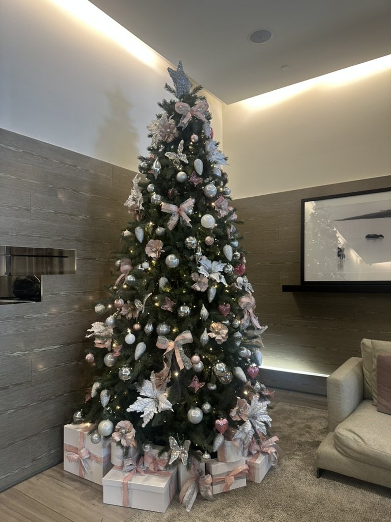 Decorated Christmas tree with pink, white, and silver ornaments, ribbons, and wrapped presents underneath in a modern living room.