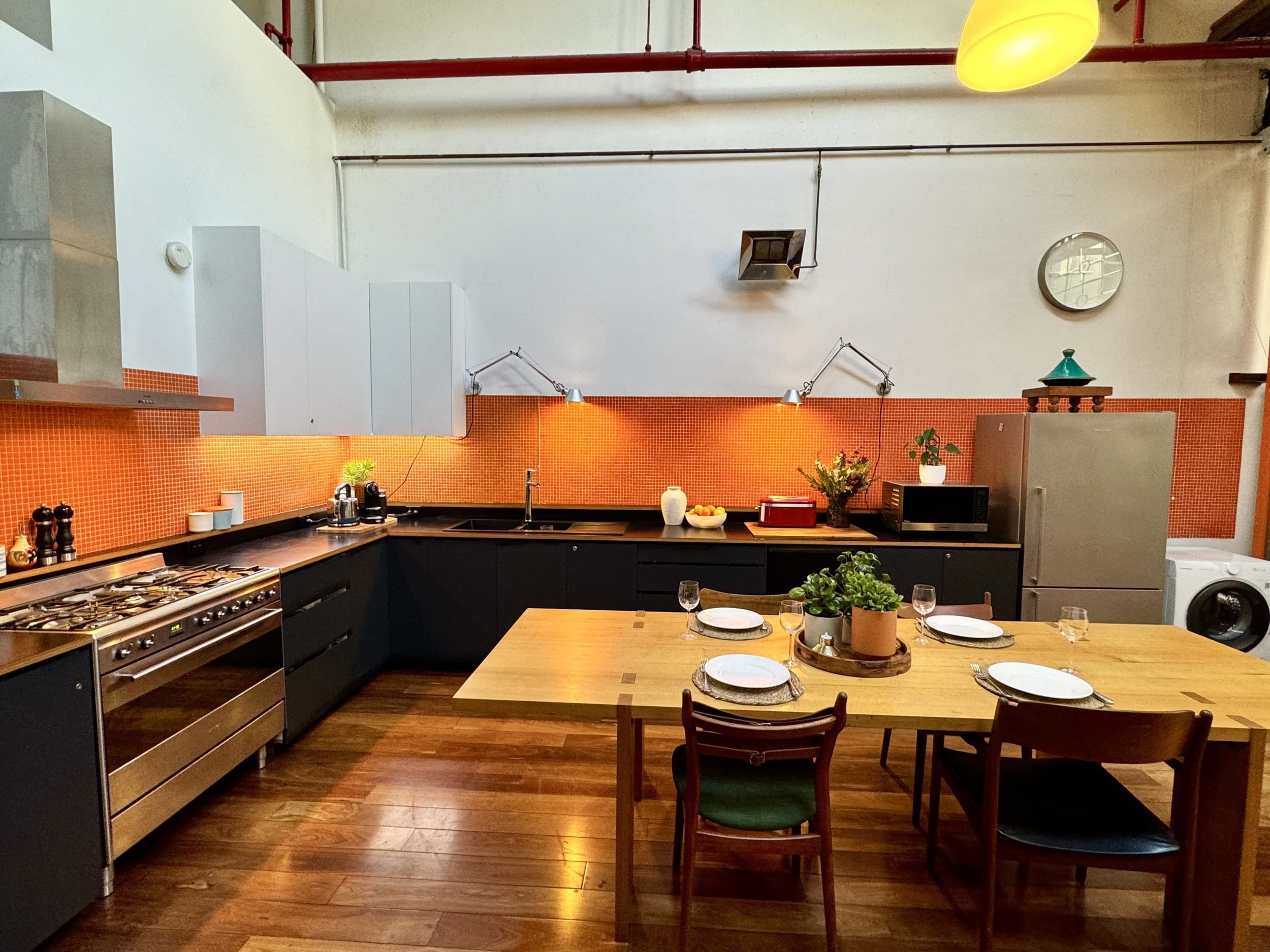 Kitchen with orange tiled backsplash, wooden dining table set with plates and glasses, black cabinets, stainless steel appliances, white walls, and yellow ceiling light.
