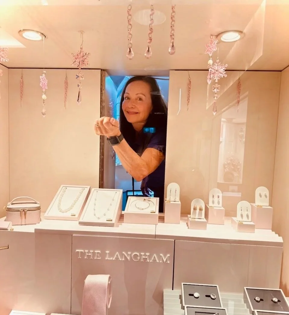 Vikki Reade from Visual Statements stands behind a jewelry display counter, smiling and looking through a mirror. The display has necklaces, earrings, and rings, with a sign that reads 'The Langham.'