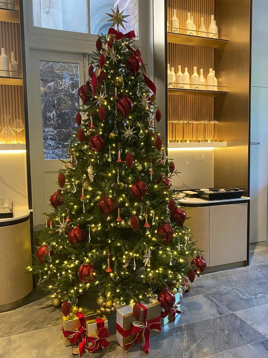 Decorated Christmas tree at Lanson Place Parliament Gardens with red, gold, and silver ornaments, surrounded by wrapped presents in red, gold, and silver ribbons, inside a modern room with glass and gold shelving.