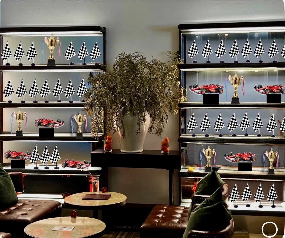 Display cases with checkered flags, trophies, and racing cars, with a large potted plant in the center and seating area in the foreground.