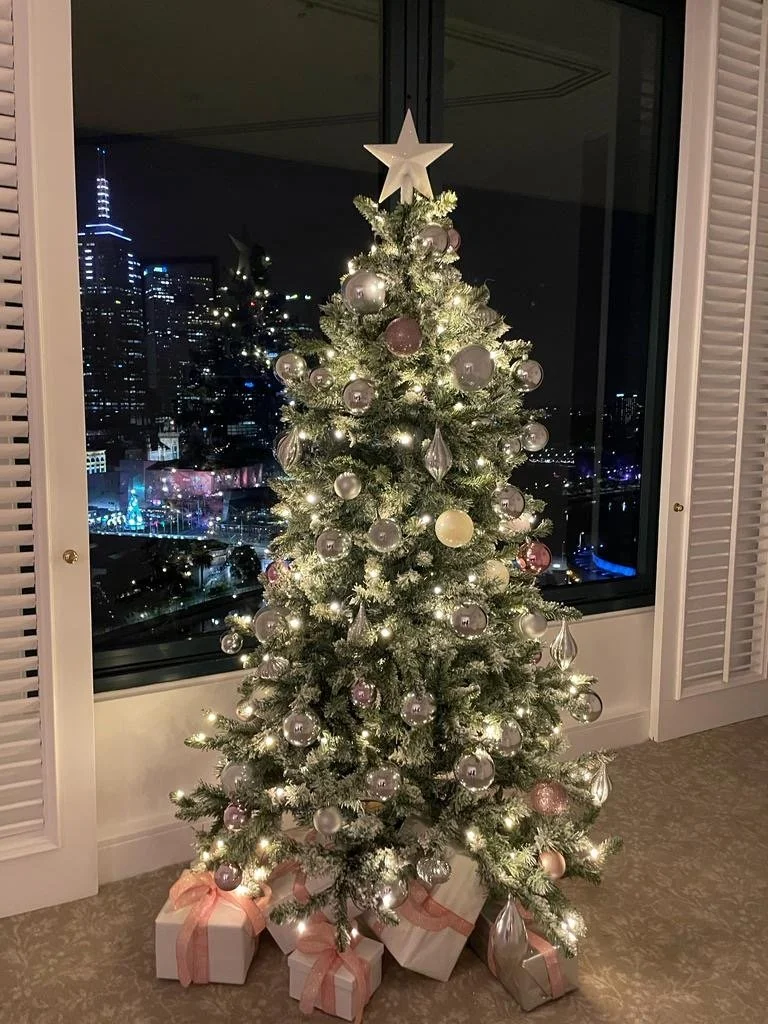 Decorated Christmas tree with white and silver ornaments, a star topper, and presents underneath, set against a large window with city lights outside at night.