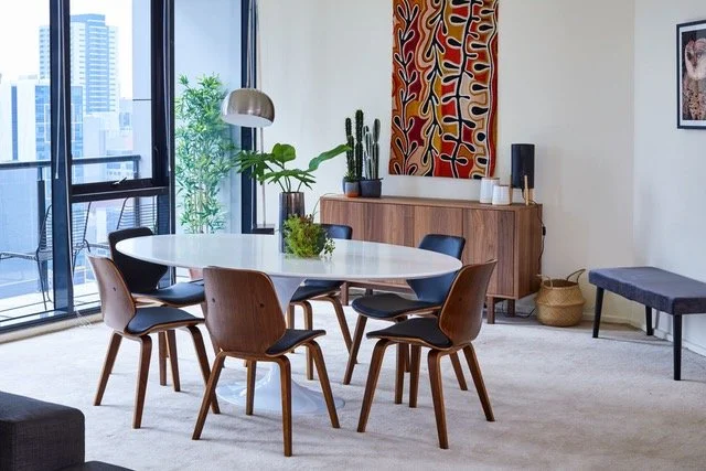 Modern dining room with a white oval table, six wooden chairs, a sideboard with plants and art, large window with city view, and contemporary decor.
