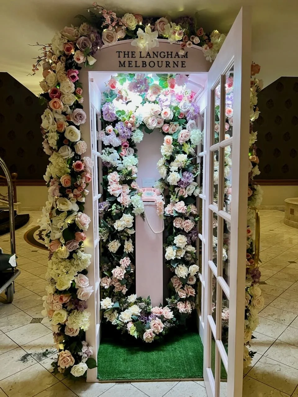 Floral arch wedding photo booth with  'The Langham Melbourne' sign and pink and white roses, lavender, and greenery, with a small green rug at the entrance.