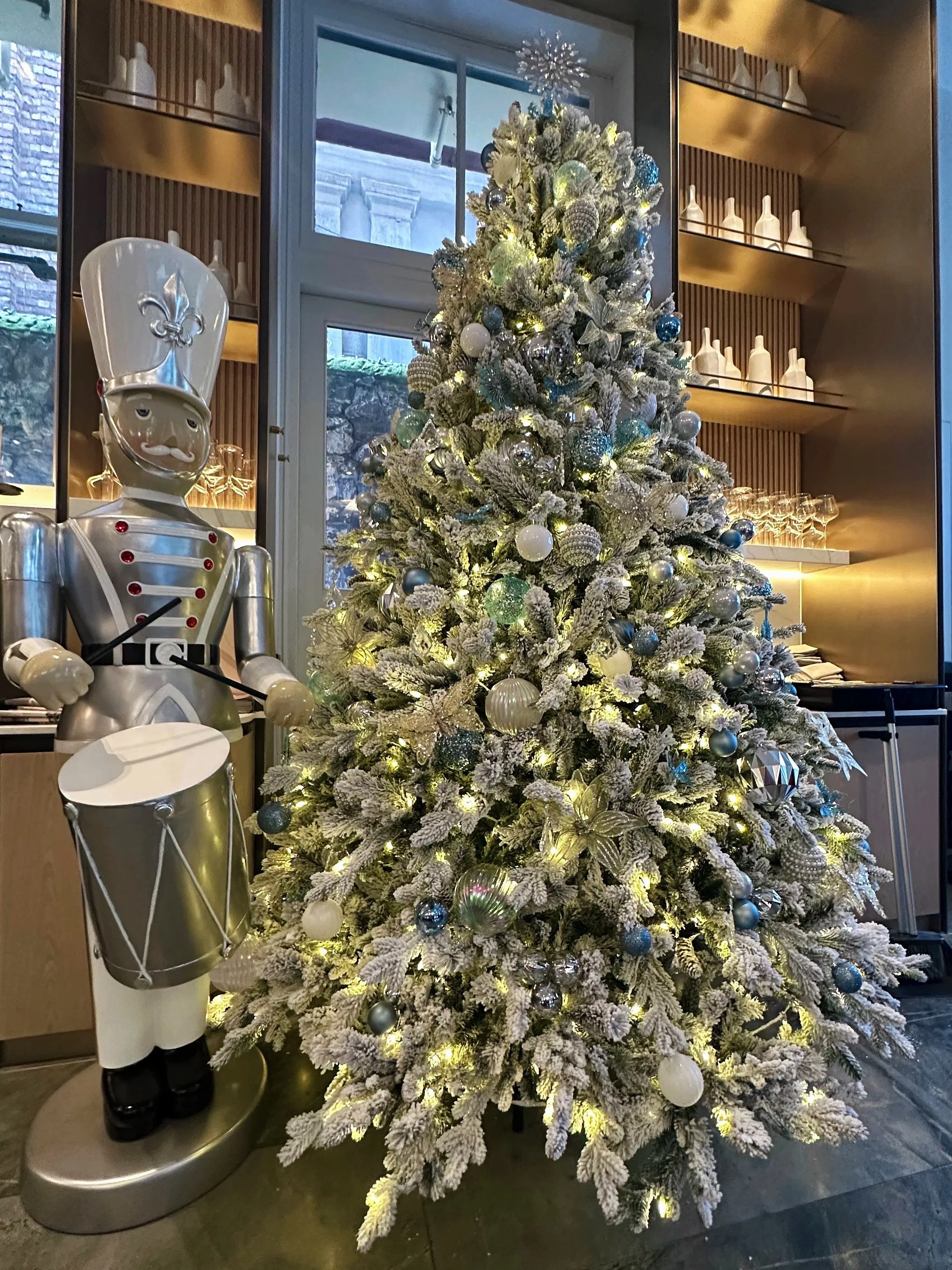 A decorated Christmas tree in Lanson Place Parliament Gardens with white, silver, and blue ornaments, illuminated with string lights, stands next to a metallic robot figure in an indoor setting.