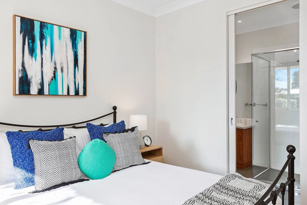 Bedroom with a bed, blue and black pillows, a round green pillow, a nightstand with a lamp and clock, abstract painting on the wall, and a glass-enclosed bathroom with a vanity inside.