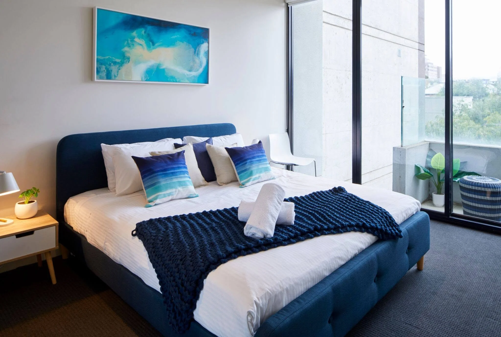 Modern bedroom with a large bed, blue headboard, and striped blue and white pillows. Nightstand with a small plant and lamp. Floor-to-ceiling windows with a balcony overlooking trees and buildings. Abstract blue and white painting on the wall.