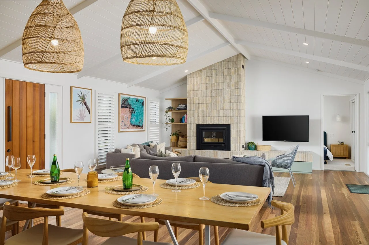 Open-concept living and dining room with wooden dining table set with plates, glasses, and bottles, gray sofas, a wood-burning fireplace, and mounted TV, decorated with framed art and wooden accents.
