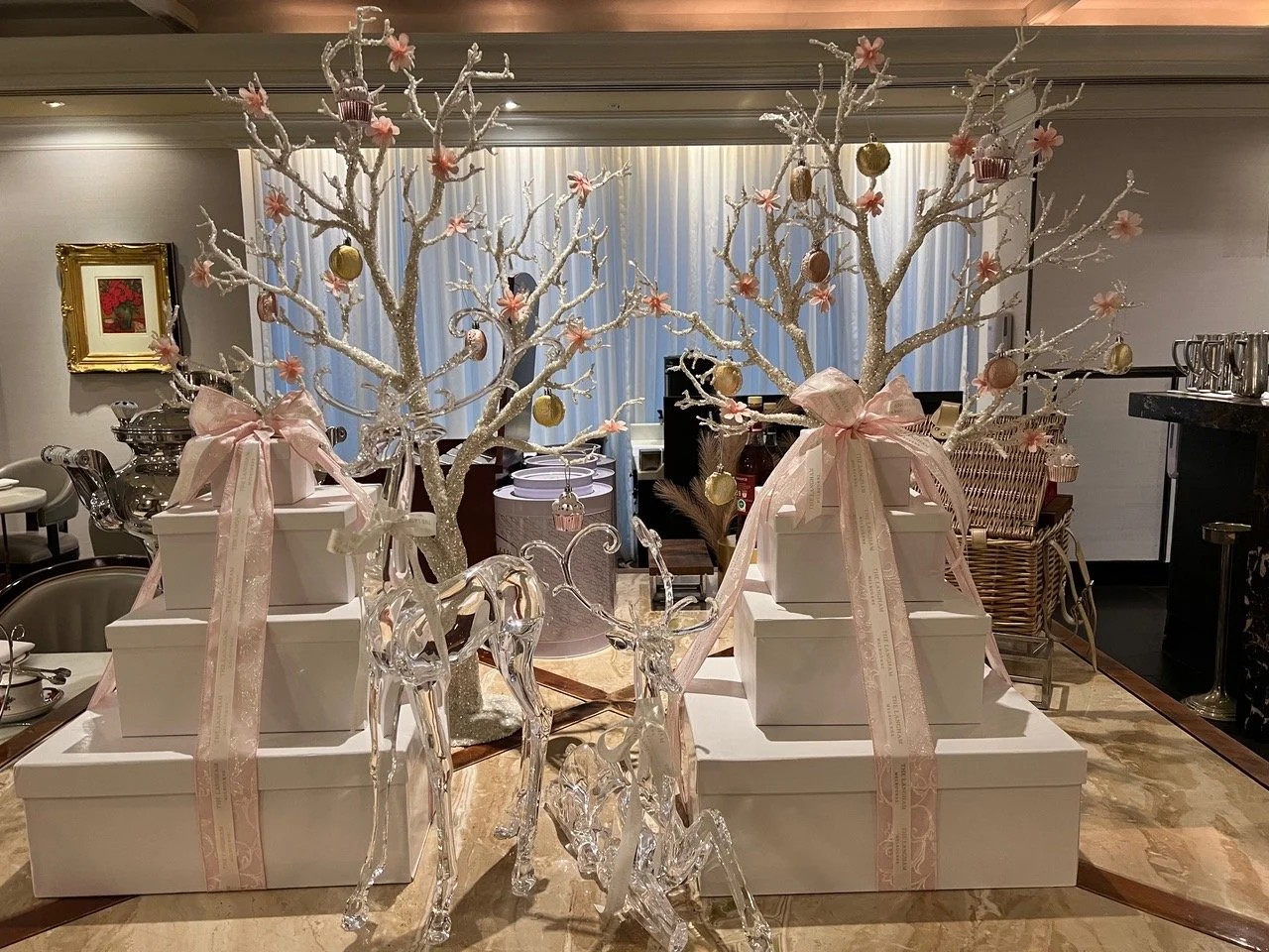Decorative display with white tiers, pink bows, and trees with ornaments and pink flowers, flanked by glass reindeer figures, in an elegant room.