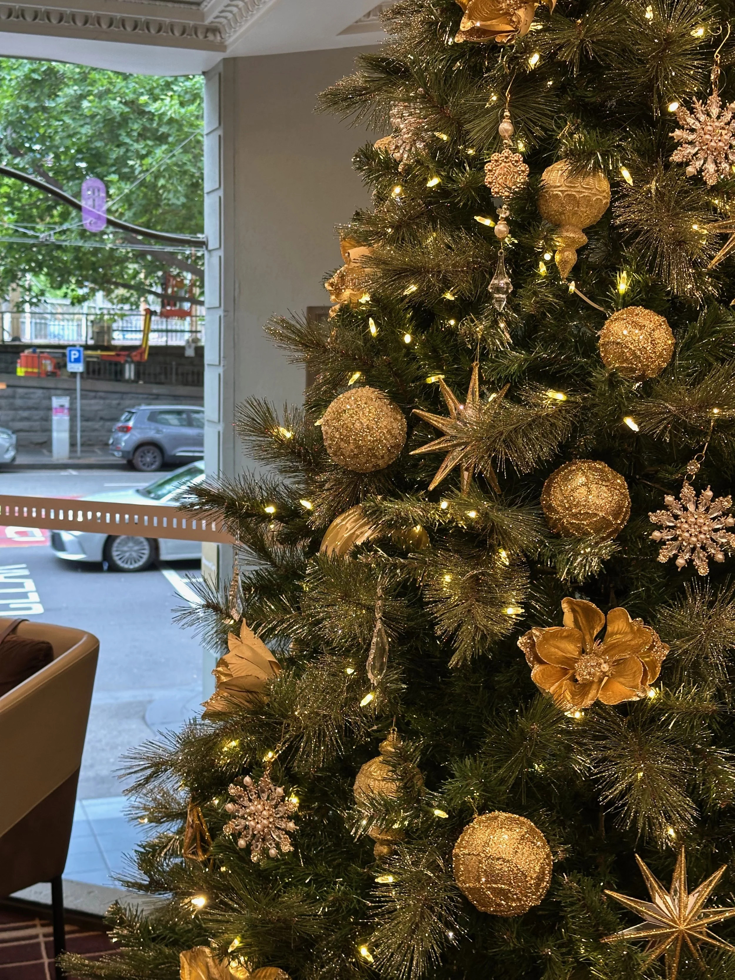 Decorated Christmas tree at RendezVous Melbourne with gold ornaments and lights inside a room, with a window showing a parking lot and street outside.