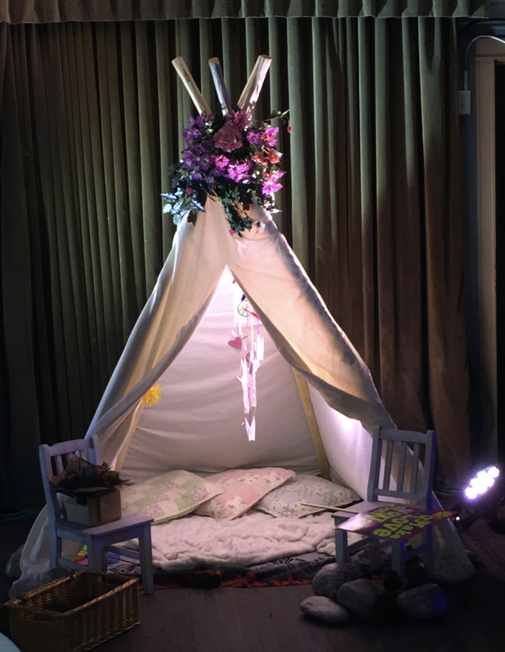A decorated teepee with flowers and lights in front of dark curtains.