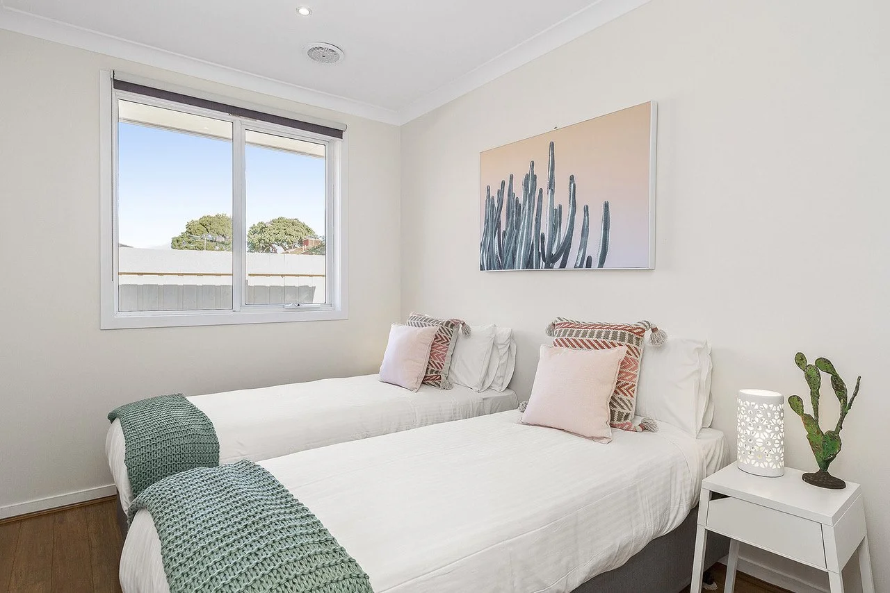 A bedroom with two single beds pushed together, with white bedding and decorative pillows. There is a window with a view of trees, a piece of artwork of cactus on the wall, a white nightstand with a modern lamp and a small decorative cactus.